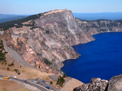 [Pink and grey cliff views at the edge of the deep blue lake. Parking lot half filled with cars is a tthe bottom of the image.]